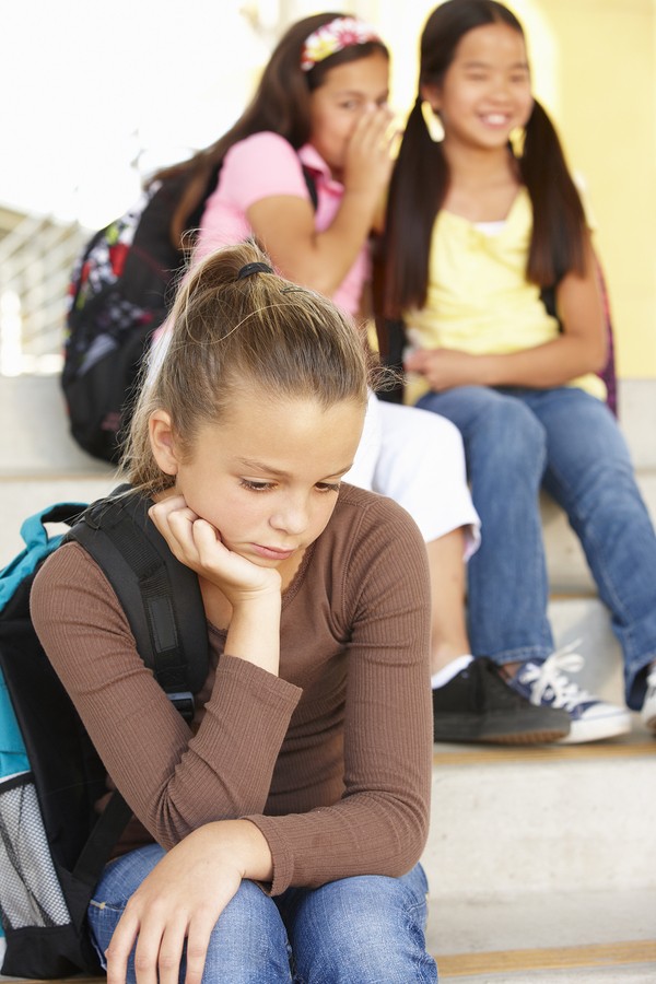 Girl in the foreground looks sad as two girls whisper behind her back.