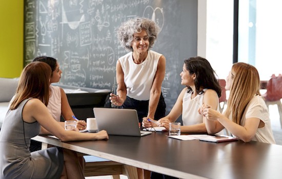 four women sitting at a table with one woman standing with a blackboard behind them